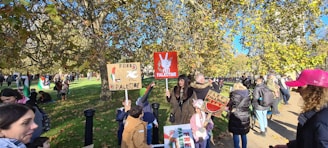 A group of people are gathered outdoors, holding signs related to Free Palestine. The scene takes place in a park-like setting with numerous trees and sunlight filtering through. Some people are wearing jackets and hats, suggesting cooler weather. There are multiple people in the background and some children among the crowd.