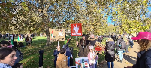 A group of people are gathered outdoors, holding signs related to Free Palestine. The scene takes place in a park-like setting with numerous trees and sunlight filtering through. Some people are wearing jackets and hats, suggesting cooler weather. There are multiple people in the background and some children among the crowd.