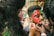 A person is having their face painted with vibrant colors, consisting of red, green, and black, in a detailed pattern. Two individuals are involved in painting, using brushes to apply makeup. Another person is holding a smartphone displaying a reference image of intricate face paint similar to the one being applied.