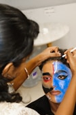 A person is having intricate face paint applied, which features bold and colorful designs. One side of the face is painted blue with striking black and white patterns, while the other side incorporates elements of red and black. The artist is focused, using a thin brush to add details.
