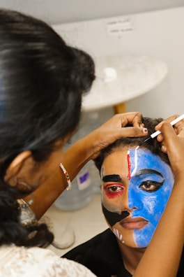 A person is having intricate face paint applied, which features bold and colorful designs. One side of the face is painted blue with striking black and white patterns, while the other side incorporates elements of red and black. The artist is focused, using a thin brush to add details.