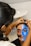 A person is having intricate face paint applied, which features bold and colorful designs. One side of the face is painted blue with striking black and white patterns, while the other side incorporates elements of red and black. The artist is focused, using a thin brush to add details.