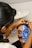 A person is having intricate face paint applied, which features bold and colorful designs. One side of the face is painted blue with striking black and white patterns, while the other side incorporates elements of red and black. The artist is focused, using a thin brush to add details.