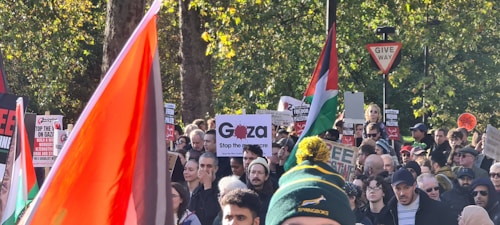 A large group of people gathered, many holding signs with messages about Gaza and Palestine. Several flags are visible, including red, white, green, and black, prominently displayed. The crowd appears dense and engaged, set against a backdrop of trees and a 'Give Way' traffic sign.