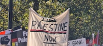 A protest scene with signs and banners, including one large beige banner with bold text reading 'Free Palestine Now.' Surrounding signs feature slogans and graphics related to the protest theme, with a backdrop of green trees visible.