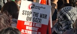 A protest scene with people gathered together, some wearing patterned scarves. A prominent sign in the foreground, associated with the Socialist Party, reads 'STOP THE WAR ON GAZA! BUILD A MASS WORKERS’ ALTERNATIVE TO WAR.'