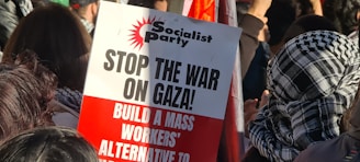 A protest scene with people gathered together, some wearing patterned scarves. A prominent sign in the foreground, associated with the Socialist Party, reads 'STOP THE WAR ON GAZA! BUILD A MASS WORKERS’ ALTERNATIVE TO WAR.'