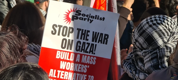 A protest scene with people gathered together, some wearing patterned scarves. A prominent sign in the foreground, associated with the Socialist Party, reads 'STOP THE WAR ON GAZA! BUILD A MASS WORKERS’ ALTERNATIVE TO WAR.'