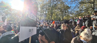 A large group of people gathered outdoors, holding signs and banners in a park setting. The crowd appears diverse and engaged in a public demonstration or protest. Sunlight filters through the trees, casting a warm glow over the scene.