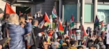 A large group of people gathered in a public space, holding Palestinian flags and signs with messages related to advocating for Palestine. The crowd appears diverse and expressive, with individuals capturing the event on phones, wearing warm clothing, and engaging with one another.
