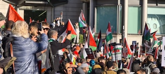 A large group of people gathered in a public space, holding Palestinian flags and signs with messages related to advocating for Palestine. The crowd appears diverse and expressive, with individuals capturing the event on phones, wearing warm clothing, and engaging with one another.