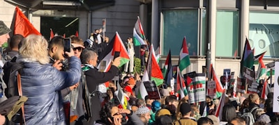 A large group of people gathered in a public space, holding Palestinian flags and signs with messages related to advocating for Palestine. The crowd appears diverse and expressive, with individuals capturing the event on phones, wearing warm clothing, and engaging with one another.