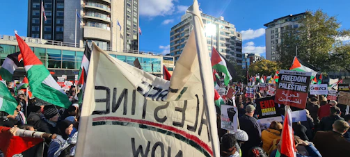 A large crowd gathers for a protest with numerous flags and banners. The scene is set in an urban environment with modern buildings in the background. People hold signs with messages, and the crowd appears to be diverse and engaged.