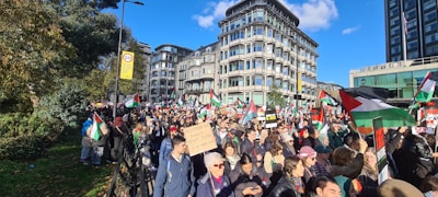 A large crowd of people is gathered for a demonstration. Many are holding flags that feature predominantly green, white, black, and red colors. Some participants carry signs with messages. The crowd is marching in what appears to be a city street, surrounded by modern buildings. The sky is clear and blue.