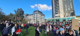 A large crowd of people gathered in an open area with several holding flags and placards. There are tall buildings in the background, along with trees and a clear blue sky. Many participants appear to be engaged in a protest or public demonstration.