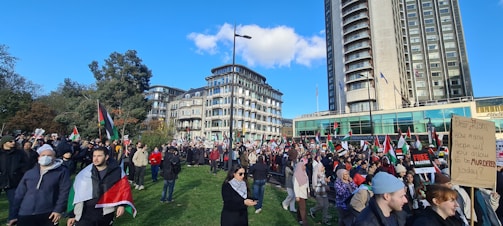 A large crowd of people gathered in an open area with several holding flags and placards. There are tall buildings in the background, along with trees and a clear blue sky. Many participants appear to be engaged in a protest or public demonstration.