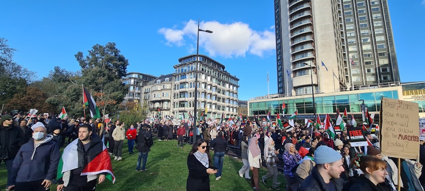 A large crowd of people gathered in an open area with several holding flags and placards. There are tall buildings in the background, along with trees and a clear blue sky. Many participants appear to be engaged in a protest or public demonstration.