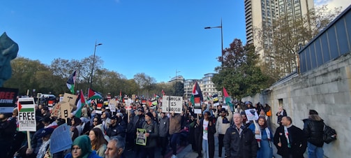 A large group of people is gathered outdoors, holding signs and banners with messages related to Palestine. Many flags are visible, and the crowd is diverse, including men and women. The setting is urban, with tall buildings and trees lining the street under a clear blue sky.
