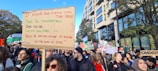 A large group of people are participating in a protest or demonstration. One person is holding a prominent cardboard sign with handwritten text. Many other participants are holding signs and banners as well. The crowd is gathered in an urban setting with trees and a modern building visible in the background.