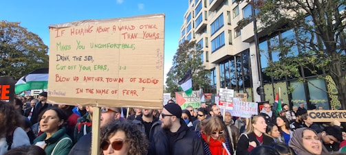 A large group of people are participating in a protest or demonstration. One person is holding a prominent cardboard sign with handwritten text. Many other participants are holding signs and banners as well. The crowd is gathered in an urban setting with trees and a modern building visible in the background.