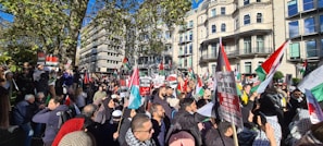 A large group of people gathers in a cityscape setting, holding various banners and flags. The demonstration is taking place in an urban area with multiple buildings in the background. Many individuals appear to be engaged in some sort of protest, with signs related to political messages.