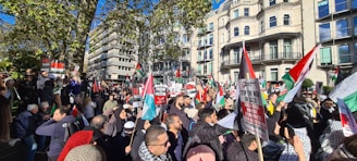 A large group of people gathers in a cityscape setting, holding various banners and flags. The demonstration is taking place in an urban area with multiple buildings in the background. Many individuals appear to be engaged in some sort of protest, with signs related to political messages.
