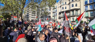 A large group of people gathers in a cityscape setting, holding various banners and flags. The demonstration is taking place in an urban area with multiple buildings in the background. Many individuals appear to be engaged in some sort of protest, with signs related to political messages.
