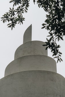 A modern, spiral-shaped architectural structure is seen from below, comprised of smooth, white concrete with a prominent fin-like extension at the top. Leaves from a nearby tree partially frame the view, adding a natural element to the scene.