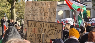 A large group of people gathering outdoors, holding signs with political messages. Some of the signs include messages supporting Palestine, with flags visible in the background. The crowd is diverse, and the setting seems to be in a public area with trees and a stone structure behind.