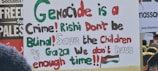 A protest sign featuring multi-colored text that reads: 'Genocide is a Crime! Rishi Don't be Blind! Save the Children of Gaza We don't have enough time!!!' The words are written in a mix of green, red, black, and brown. In the background, there are other protest signs partly visible.