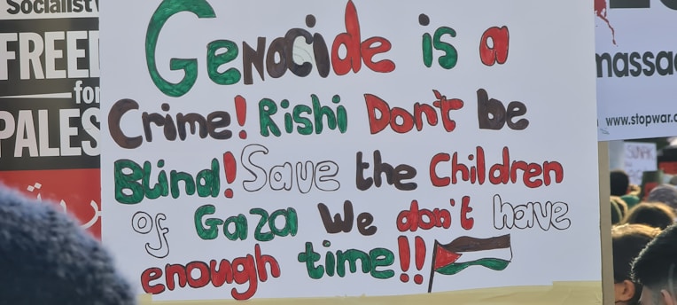A protest sign featuring multi-colored text that reads: 'Genocide is a Crime! Rishi Don't be Blind! Save the Children of Gaza We don't have enough time!!!' The words are written in a mix of green, red, black, and brown. In the background, there are other protest signs partly visible.