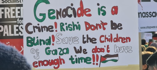 A protest sign featuring multi-colored text that reads: 'Genocide is a Crime! Rishi Don't be Blind! Save the Children of Gaza We don't have enough time!!!' The words are written in a mix of green, red, black, and brown. In the background, there are other protest signs partly visible.