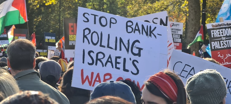 A large group of people are gathered, holding various protest signs. One prominent sign in the center reads 'Stop Bankrolling Israel's War Crimes'. Other signs include messages supporting Palestine and criticizing Israeli actions. There are flags and a mixture of people, indicating a protest or demonstration.