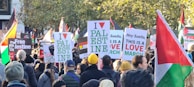 A large group of people are gathered for a protest or march, holding various signs and banners expressing support for Palestine. Numerous Palestinian flags can be seen throughout the crowd, and the signs display messages advocating for freedom and resistance.