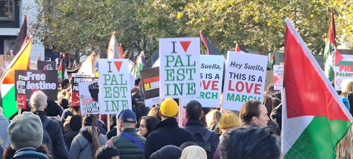 A large group of people are gathered for a protest or march, holding various signs and banners expressing support for Palestine. Numerous Palestinian flags can be seen throughout the crowd, and the signs display messages advocating for freedom and resistance.