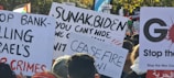 A collection of protest signs is held up in a crowd. Messages on the signs include demands for ceasefire, criticisms of political leaders, and calls to stop war crimes. The background includes trees and more protest signs, some with political and peace-related messages.