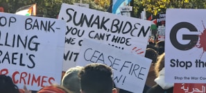 A collection of protest signs is held up in a crowd. Messages on the signs include demands for ceasefire, criticisms of political leaders, and calls to stop war crimes. The background includes trees and more protest signs, some with political and peace-related messages.