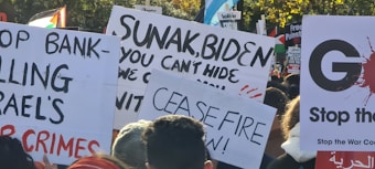 A collection of protest signs is held up in a crowd. Messages on the signs include demands for ceasefire, criticisms of political leaders, and calls to stop war crimes. The background includes trees and more protest signs, some with political and peace-related messages.