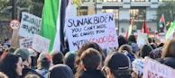 A crowded protest scene with many people holding up signs and banners. Prominent signs include phrases like 'Ceasefire Now!' and 'You Can't Hide, We Charge You With Genocide'. Flags and various protest placards are visible in the background.