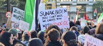 A crowded protest scene with many people holding up signs and banners. Prominent signs include phrases like 'Ceasefire Now!' and 'You Can't Hide, We Charge You With Genocide'. Flags and various protest placards are visible in the background.