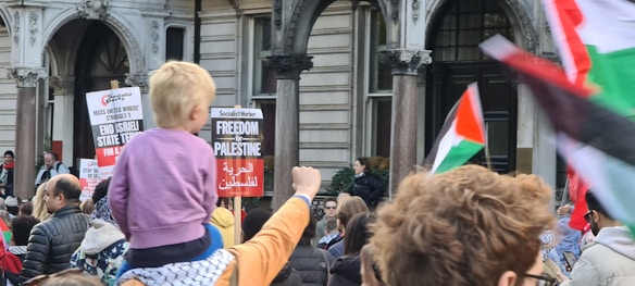 A large group of people gathered in front of a historic building, holding signs and flags. One prominent red, white, green, and black flag is visible among others. A child sits on an adult's shoulders, observing the scene. The signs carry messages related to political protest and solidarity.