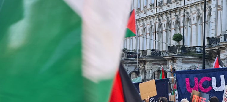 A crowd of people is gathered in front of a historical building with ornate architecture. Several signs and flags are visible, including a prominent green, white, and red flag. The gathering appears to be a rally or protest with banners being held up by participants.