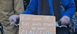 A person wearing a blue jacket and purple gloves is holding a cardboard sign with handwritten text. The text criticizes political leaders and calls for a ceasefire. The background shows other people partially visible.