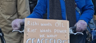 A person wearing a blue jacket and purple gloves is holding a cardboard sign with handwritten text. The text criticizes political leaders and calls for a ceasefire. The background shows other people partially visible.