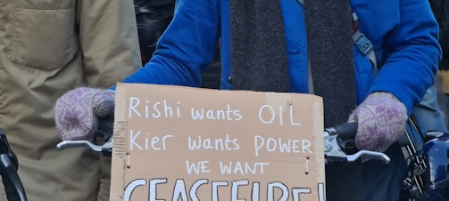 A person wearing a blue jacket and purple gloves is holding a cardboard sign with handwritten text. The text criticizes political leaders and calls for a ceasefire. The background shows other people partially visible.