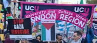 A crowded protest scene with people holding various signs and banners. A large banner reads 'UCU University and College Union London Region' with a background illustration of domed buildings. Other signs include messages calling for ceasefire and stopping the Gaza genocide.