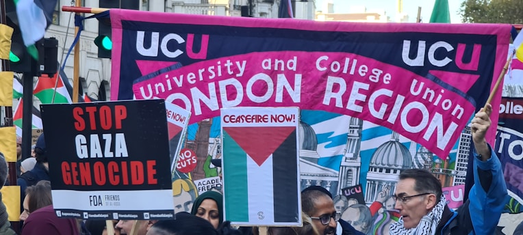 A crowded protest scene with people holding various signs and banners. A large banner reads 'UCU University and College Union London Region' with a background illustration of domed buildings. Other signs include messages calling for ceasefire and stopping the Gaza genocide.