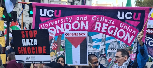 A crowded protest scene with people holding various signs and banners. A large banner reads 'UCU University and College Union London Region' with a background illustration of domed buildings. Other signs include messages calling for ceasefire and stopping the Gaza genocide.