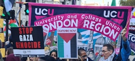 A crowded protest scene with people holding various signs and banners. A large banner reads 'UCU University and College Union London Region' with a background illustration of domed buildings. Other signs include messages calling for ceasefire and stopping the Gaza genocide.