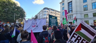 A large crowd gathers on a street for a protest or demonstration. They are holding various signs and banners, including a prominent one displaying 'UCL UCU' and others with phrases related to a campaign. Many participants hold flags and placards, and the scene is set against an urban backdrop with tall buildings and trees in the distance.